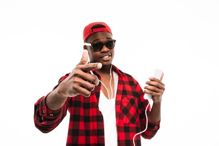 Handsome african young man listening to music from mobile phone and pointing on you over white...