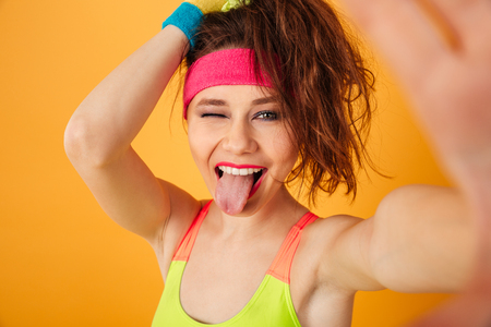 Cheerful young sportswoman making selfie, winking and showing tongue over yellow backgroundの写真素材