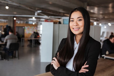 Happy asian young businesswoman standing with arms crossed in officeの写真素材