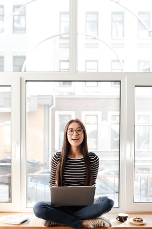 Vertical image of Happy Asian woman in eyeglasses which sitting on windowsill with laptop and looking at cameraの写真素材