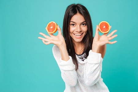 Happy beautiful young woman standing and holding two halves of grapefruit over blue background