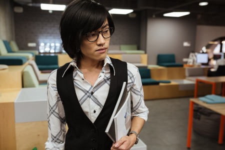 Image of attractive young asian man wearing glasses dressed in jacket and shirt standing indoors and...