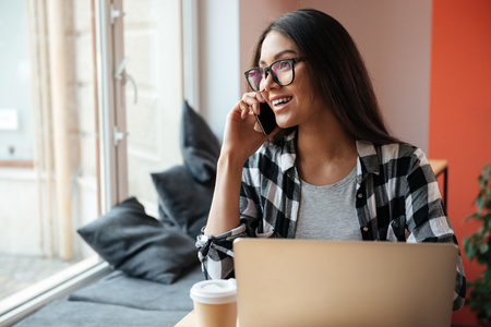 Picture of amazing young caucasian woman wearing glasses sitting indoors using laptop computer while talking by phone and drinking coffee. Coworking concept.の写真素材