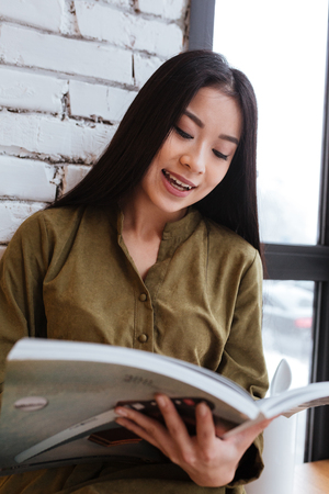 Image of a cheerful asian young lady sitting at the cafe near window while reading magazine. Looking aside.の写真素材
