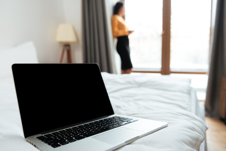 Close up portrait of a blank screen laptop laying on bed with businesswoman standing at the window on backgroundの写真素材