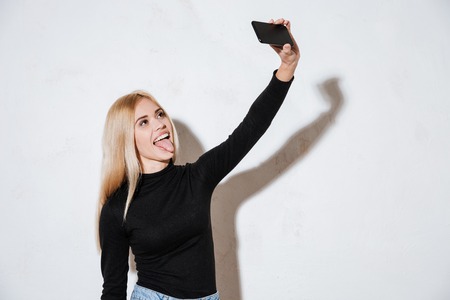 Smiling funny girl showing tongue while taking selfie isolated on a white background