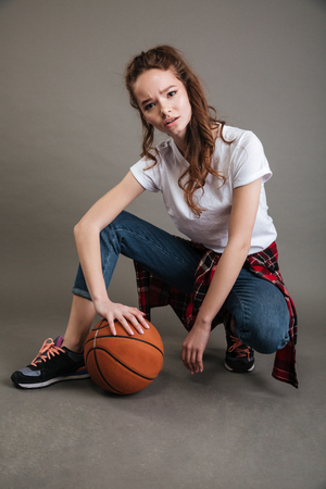 Portrait of a pretty casual teenage girl sitting and posing with basketball ball isolated on the gray backgroundの写真素材