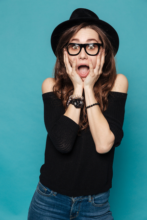 Surprised young woman in hat standing and looking at camera with hands on her face over blue backgroundの写真素材