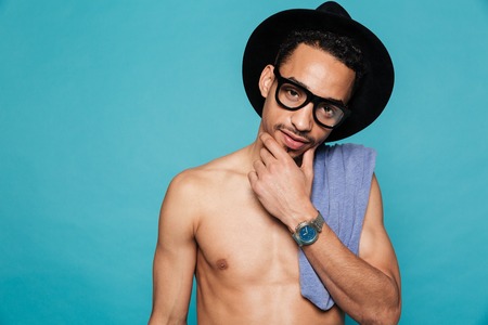 Portrait of a young casual shirtless african man in hat and eyeglasses thinking isolated on the blue...