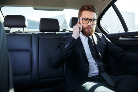 Portrait of an angry frustrated business man in eyeglasses talking on mobile phone while sitting in the back seat of carの写真素材