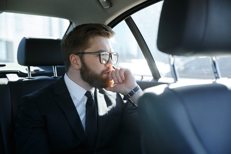 Potrait of a concentrated bearded business man in eyeglasses sitting in the back seat of car