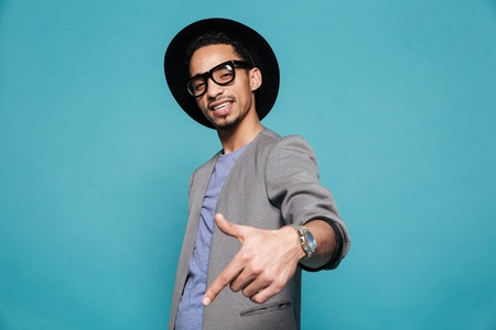 Portrait of a confident afro american man in eyeglasses and hat pointing finger at copyspace over blue backgroundの写真素材