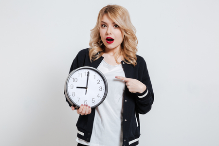 Portrait of a surprised young teenager girl holding alarm clock and pointing finger isolated on a white backgroundの写真素材