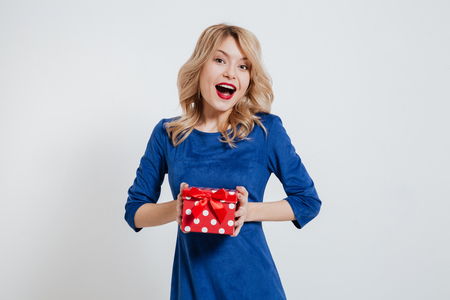 Picture of cheerful young woman dressed in blue dress holding gift box over white background.の写真素材