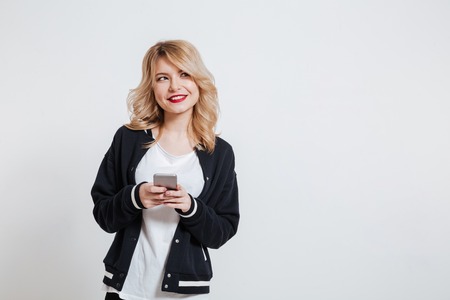 Portrait of a pretty young girl in casual clothes holding mobile phone and looking away isolated on the white backgroundの写真素材