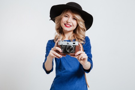 Picture of pretty young woman dressed in blue dress wearing hat holding camera and posing over white...