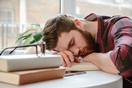 Image of sleeping bearded young man student lies on hands in cafe near books and glasses. Eyes...