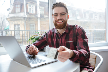 Photo of cheerful bearded young man sitting in cafe while using laptop computer and showing debit card to camera.の写真素材