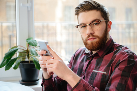 Image of handsome bearded young man student wearing eyeglasses sitting in cafe while using mobile phone.の写真素材