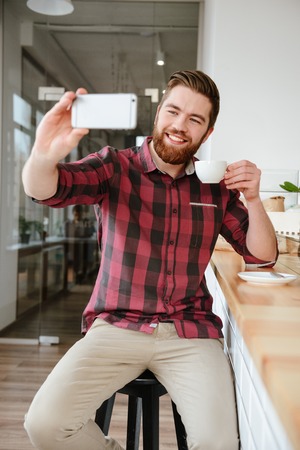Young bearded man student taking selfie and holding cup of coffee while sitting at cafe