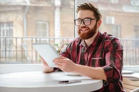 Picture of happy bearded young man student wearing glasses sitting in cafe while using tablet computer. Looking aside.の写真素材