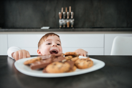 Photo of pretty boy standing in kitchen while tries to take cookies and chocolate.の写真素材