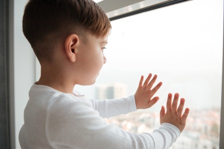 Photo of alone little boy near window waiting for parents at home. Look at window.