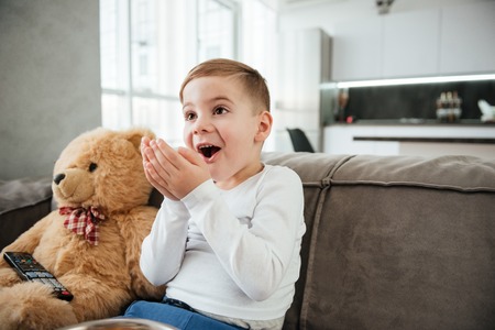 Picture of surprised boy on sofa with teddy bear at home watching TV while eating chips.の写真素材