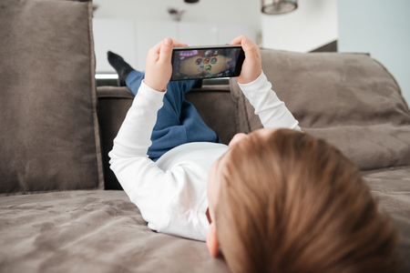 Back view picture of little boy holding smartphone while lies on sofa at home. Look at phone.の写真素材