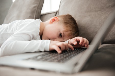 Picture of little alone sad boy lies on sofa while using laptop computer.