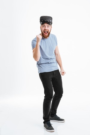 Young happy bearded man wearing virtual reality device standing over white background and looking at...