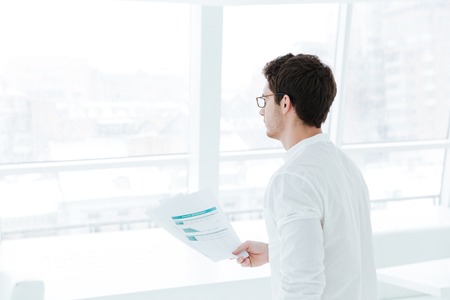 Photo of young man dressed in white shirt holding documents near big white window.の写真素材