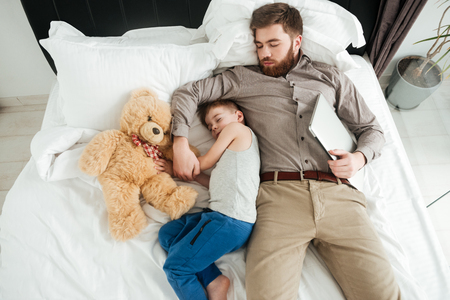 Image of cute little boy sleeping at home in bed with his bearded father and toy.の写真素材