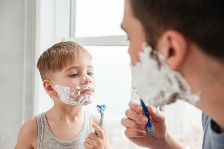 Image of funny father and son are applying shaving foam on their faces and smiling while shaving in bathroomの写真素材