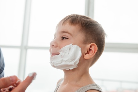 Father and son are applying shaving foam on their faces and smiling while shaving in bathroom