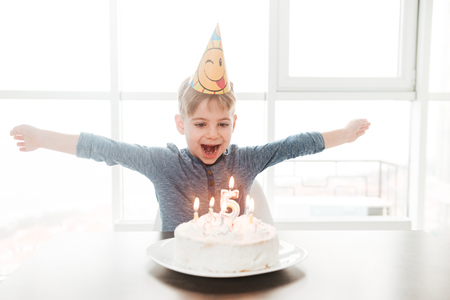 Picture of cheerful birthday boy sitting in kitchen near cake while smiling. Look at cake while...