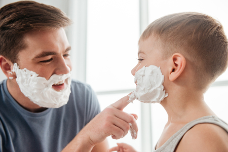 Picture of handsome father and son are applying shaving foam on their faces and smiling while shaving in bathroomの写真素材