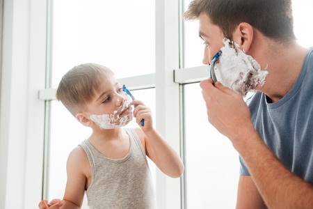 Photo of young happy father and son are applying shaving foam on their faces and smiling while shaving in bathroomの写真素材