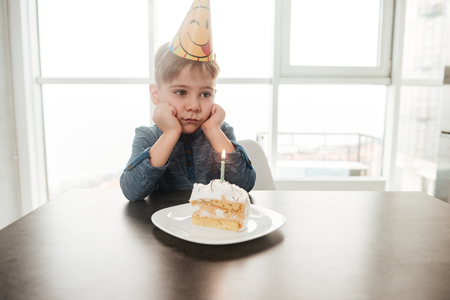 Image of little sad birthday boy sitting in kitchen near cake alone. Look aside.の写真素材