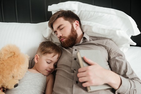 Image of boy sleeping at home in bed near toy with his bearded father holding book.の写真素材