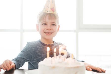 Photo of little cute happy birthday boy sitting in kitchen near cake while smiling. Look aside.の写真素材