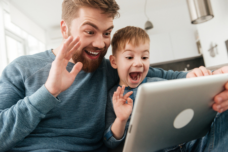 Image of cheerful father talking with friends by tablet computer and waving with his little cute son.の写真素材