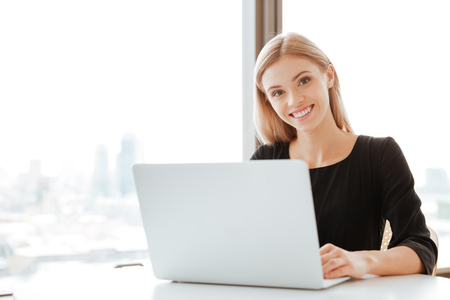 Picture of cheerful young lady worker sitting in office while using laptop computer and typing by keyboard. Look at camera.の写真素材