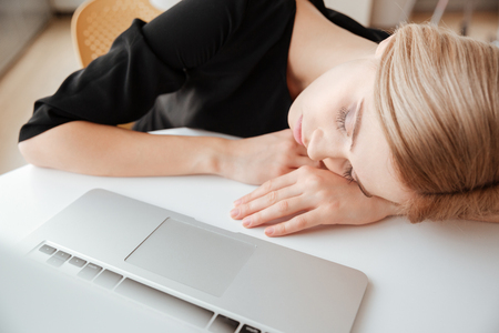 Image of young tired lady worker lies on table in office near laptop computer and sleeping. Eyes closed.の写真素材