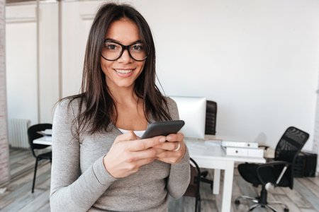 Picture of pretty smiling lady wearing eyeglasses standing in office while using telephone. Look at camera.の写真素材