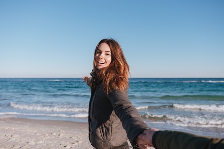 First person view of man holding hand of smiling woman in warm jacket on the beachの写真素材