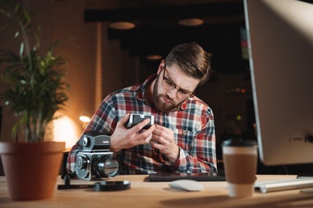 Young bearded man in plaid shirt trying to fix old camera while sitting at his workplace