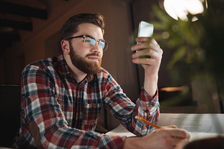 Smiling young man using his smartphone while sitting at his working placeの写真素材