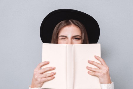 Close up portrait of a young woman in hat covering her mouth with book and winking isolated on the gray backgroundの写真素材