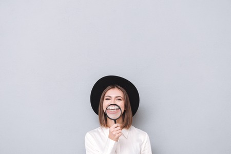 Portrait of a young woman looking at camera and having fun with magnifying glass over gray...